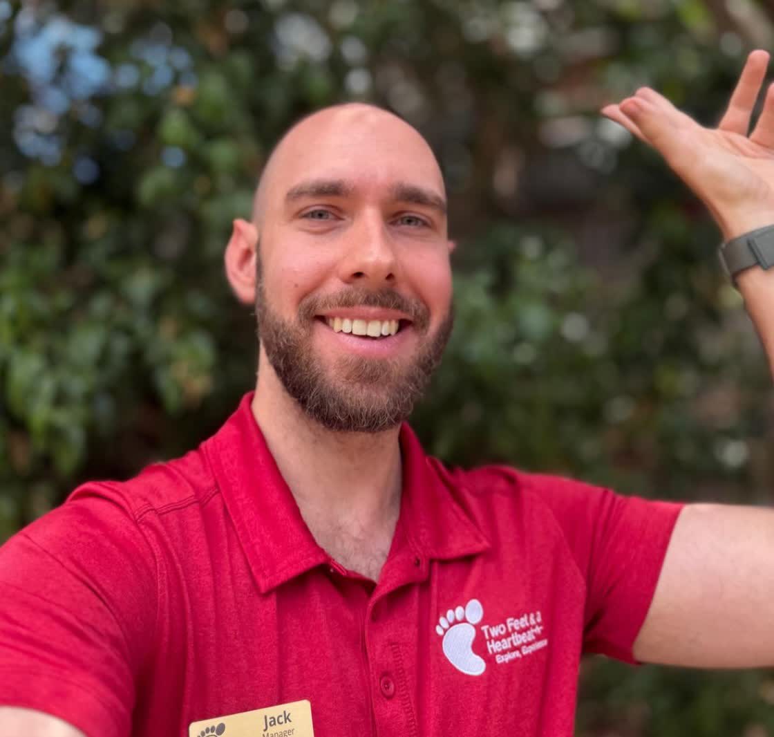 Two Feet guide Jack waving in his red team polo and name badge in front of leafy greenery