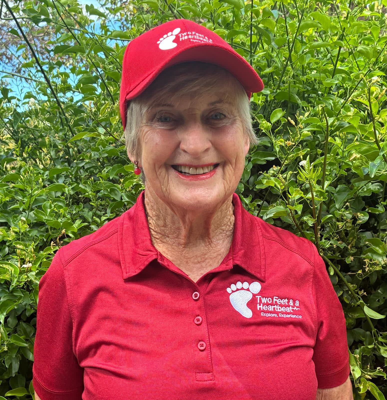 Two Feet guide Joni in the red team cap and polo, smiling in front of a jasmine hedge